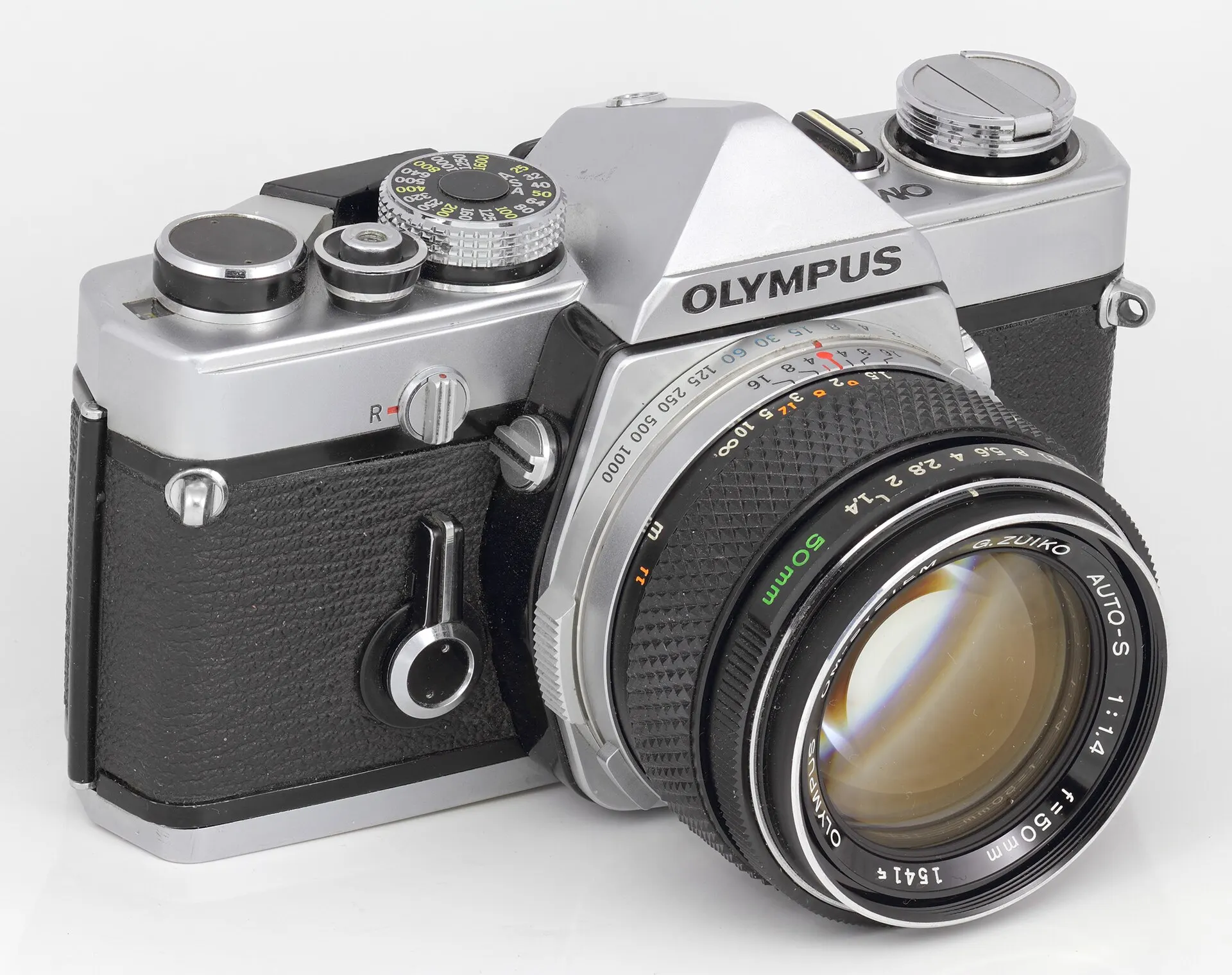 Chrome Olympus OM-1 body from 1973-1974 with a G.Zuiko Auto-S 50mm f/1.4 lens, seen from the front left on a light background