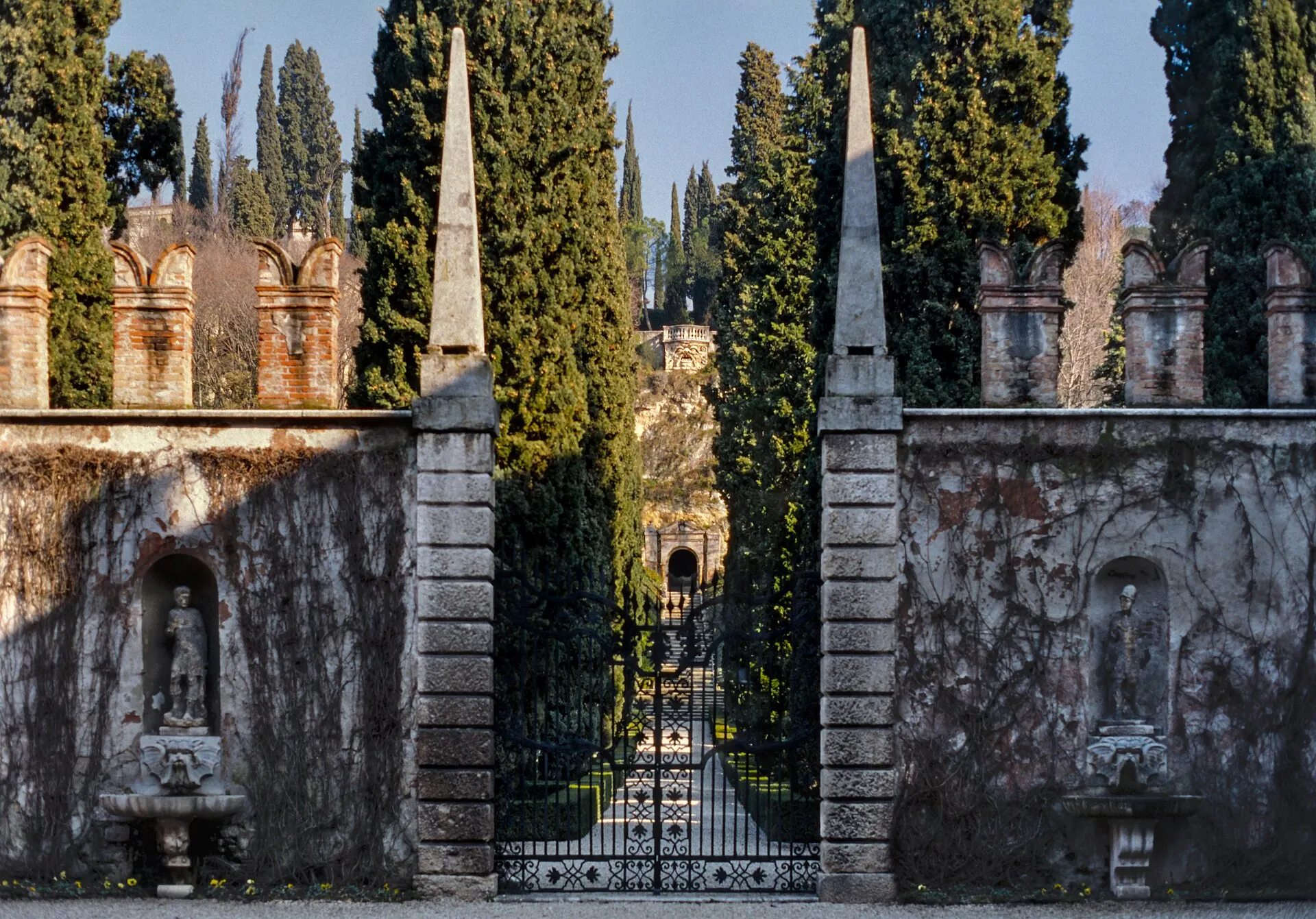 Main gate of a formal Italian garden with two obelisks and an avenue of tall cypresses behind it