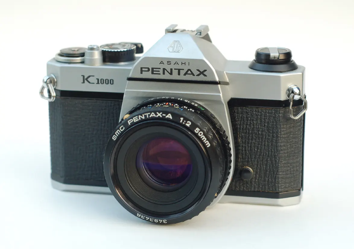 Silver and black Pentax K1000 with an SMC Pentax-A 50mm f/2 lens on a plain white background