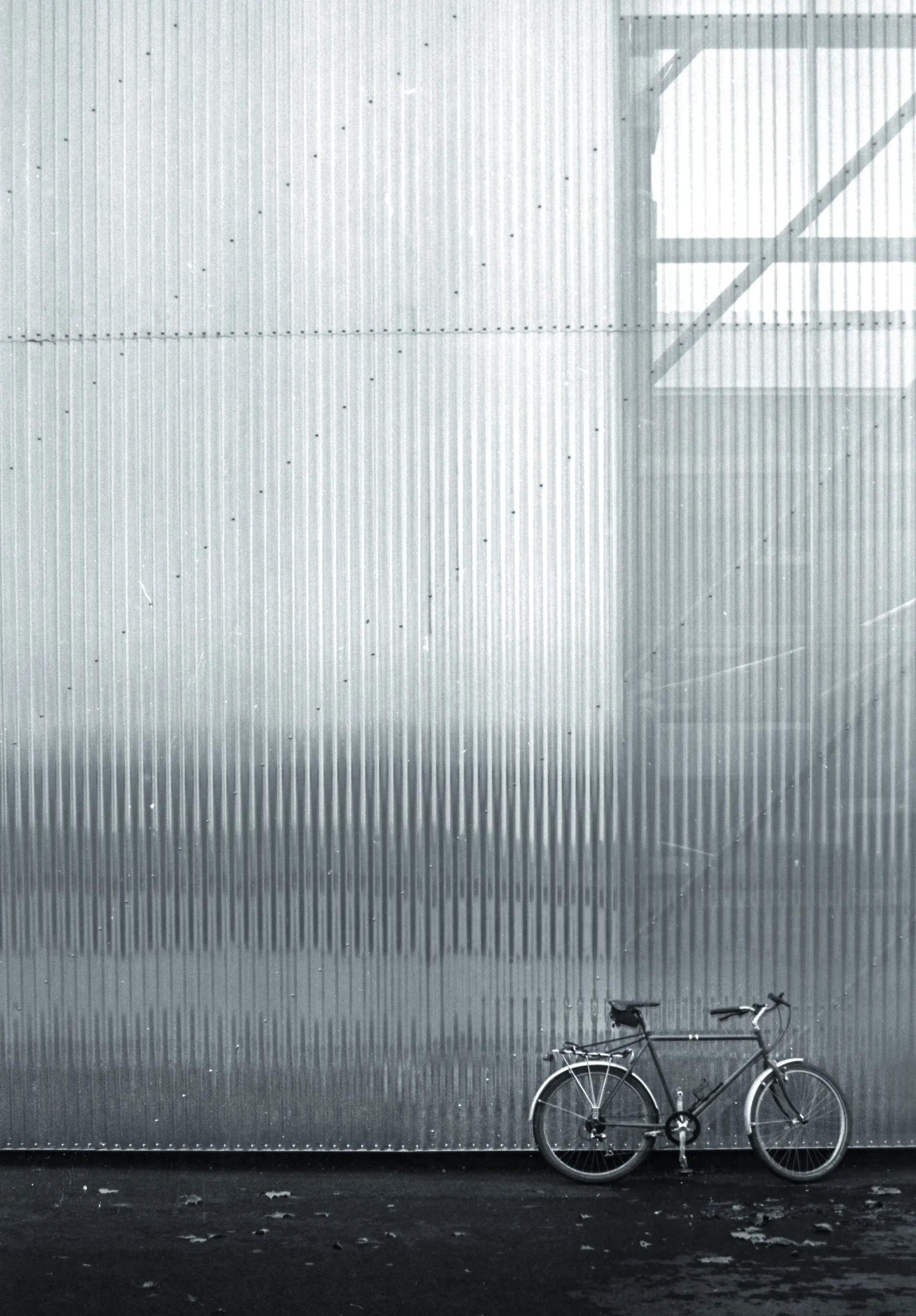 Bicycle parked against a tall corrugated hangar wall, in black and white