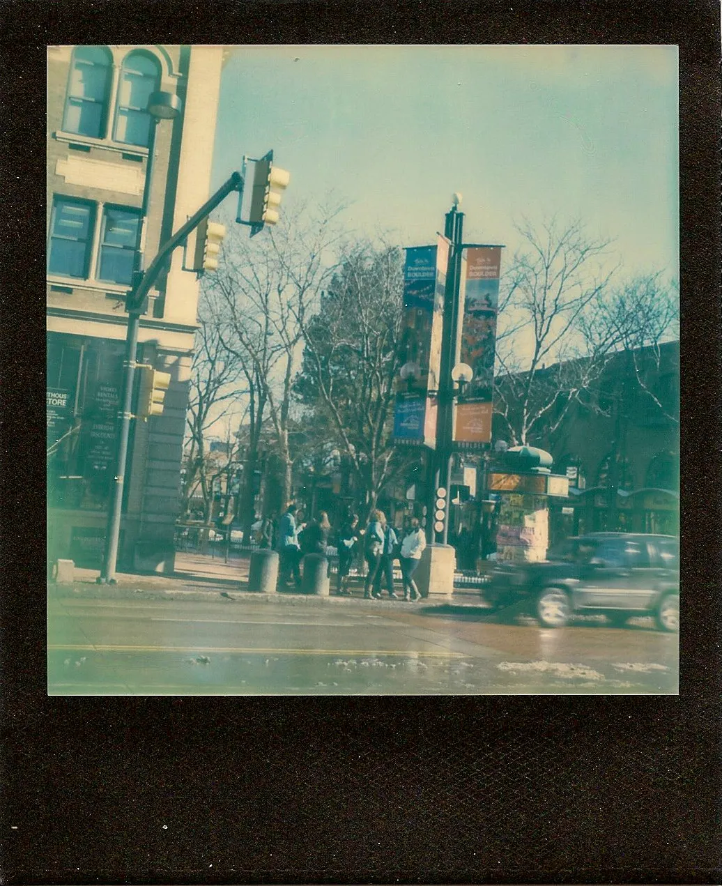 Polaroid print of a downtown street corner with traffic lights, bare trees and Downtown Boulder banners on a lamppost, in soft, cool color