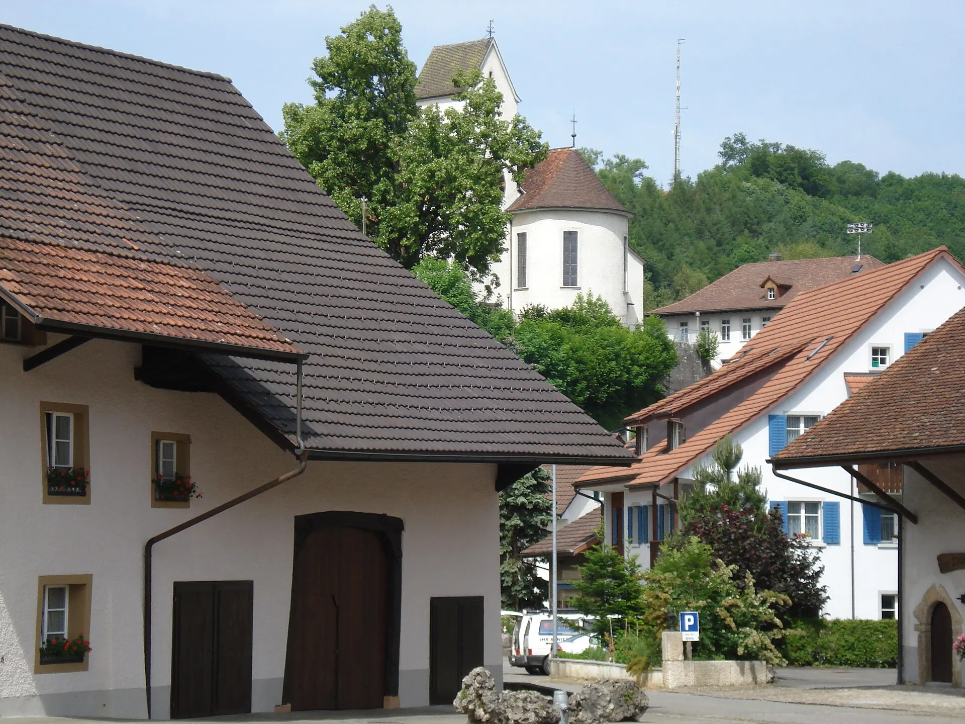 Old houses with tiled roofs around a village square, a church on a wooded hill behind